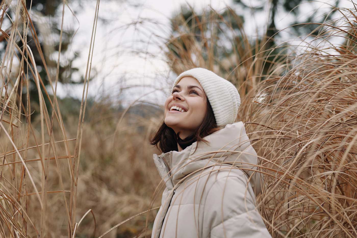 Woman outdoors smiling in a light jacket among tall grass, enjoying a crisp day. A candid portrait in nature with soft earth tones, a calm mood and tranquil scenery.